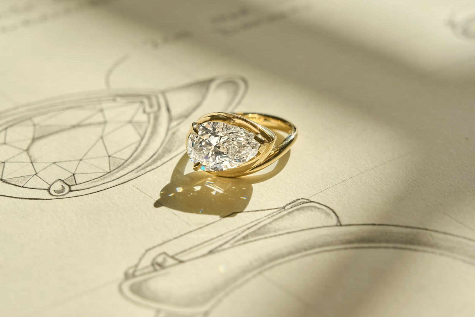 A gold-tone Frank Darling ring with a pear-shaped center stone resting on pencil drawings of ring settings.